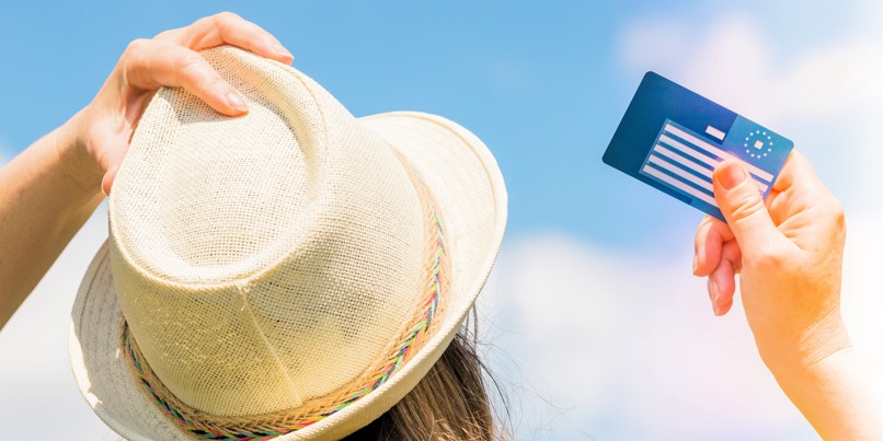 Kvinde med solhat holder EU-sygesikringskort op mod blå himmel